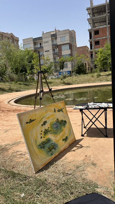 An outdoor painting setup with an easel displaying a yellow abstract artwork, positioned near a pond with buildings under construction visible in the background.