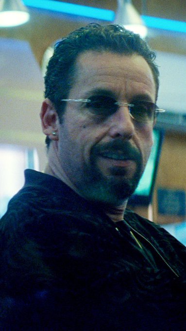 A close-up portrait of a man with short dark hair, a goatee, and rimless glasses wearing a dark jacket.