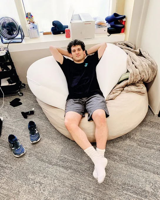 A young man relaxes on a large white bean bag chair in a casual indoor setting, with sneakers and game controllers scattered on the carpet nearby.