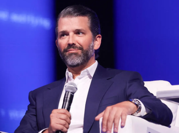 Donald Trump Jr. speaks into a microphone while seated on a white chair against a blue backdrop, appearing to be at a public event or conference. The image accompanies commentary about his significant $300 million investment in Polymarket, highlighting the irony of his financial stake in prediction markets while his father's administration simultaneously battles state-level regulation efforts of those same platforms.