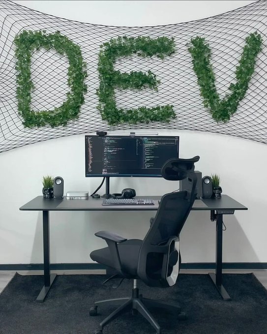 A developer desk setup featuring a monitor with code, a keyboard, and a black ergonomic chair, with the word 'DEV' spelled out in green artificial ivy on a black mesh wall above.