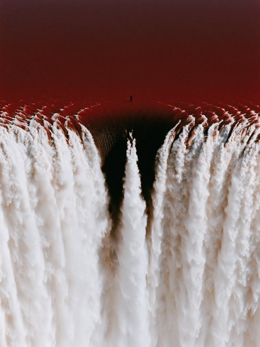 An aerial, surreal view shows a solitary figure standing at the edge of a massive, deep crater filled with jagged, white, ice-like formations against a dark red background.
