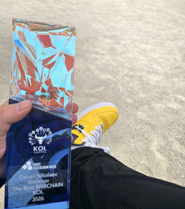A person holds a holographic "KOL Capital" award from Paris Blockchain Week, inscribed to "Danail Nikolaev Stefanov" as "The Best BNBCHAIN KOL 2026," showcasing their yellow and white sneakers in the background while proudly displaying the recognition tied to their post about BNBCHAIN's achievements and support for builders.