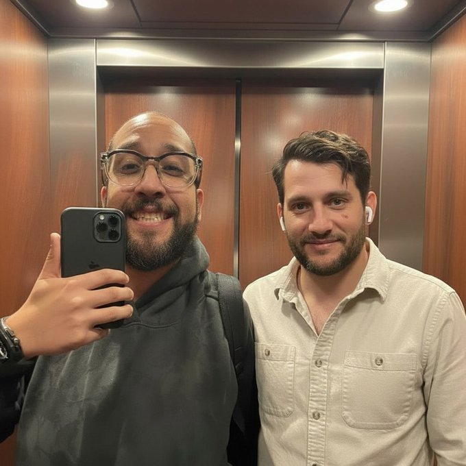 Two men take a mirror selfie inside an elevator, with the man on the left holding up an iPhone to capture the shot while both smile at the camera, fitting the upbeat "GM" (good morning) vibe of the post.