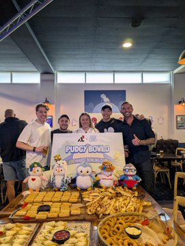 A group of five people pose behind a table displaying an assortment of pastries and plush toys, with a sign in front reading "Pudgy Bowls - Dessert Heaven."