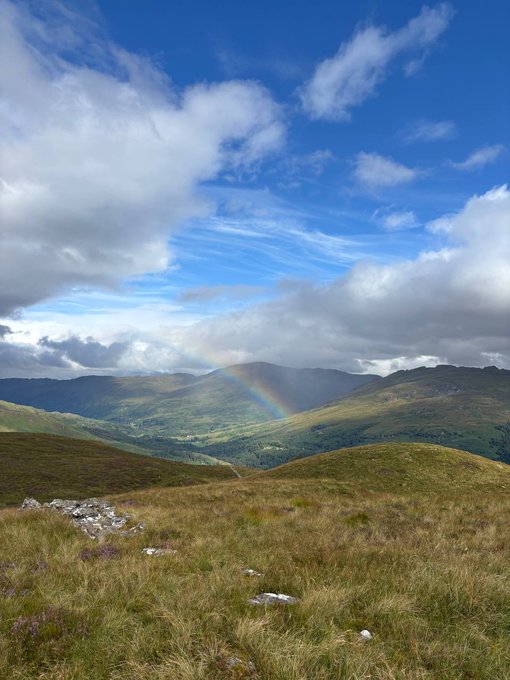 A scenic view from a Scottish hillside featuring a vibrant rainbow arching over misty, rolling green mountains under a partly cloudy sky, perfectly capturing the natural beauty celebrated in the team's "Greetings from our Scotland offsite" message.