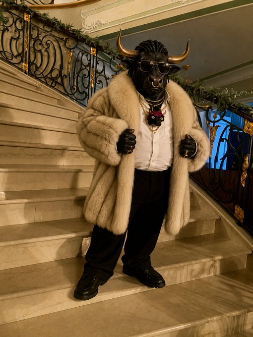 A person wearing a bull mask, sunglasses, and a large fur coat stands on a marble staircase, accessorized with heavy gold chains.