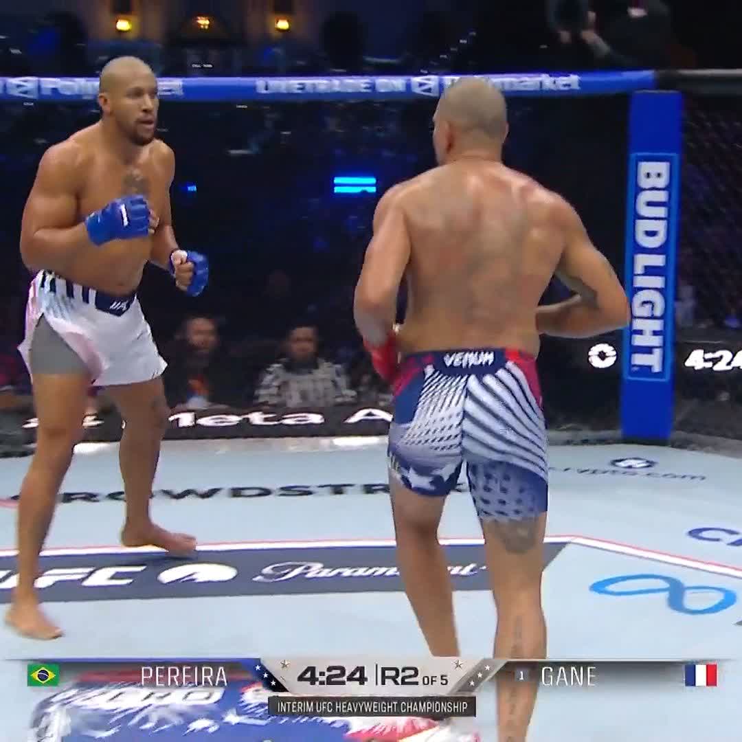 The image shows two men fighting each other, with one man wearing a blue and white striped pair of shorts and the other wearing an American flag shorts. The fighters are both barefoot, suggesting that they might be at a professional wrestling event or a similar type of combat sport. The fighter on the left is holding his opponent's arm in front of him, indicating that he has just landed a punch. The image also includes text that reads "GANE WITH MASSIVE MOMENT IN ROUND TWO #UFCWhiteHouse [ Title fights presented by @Cryptocom ]".