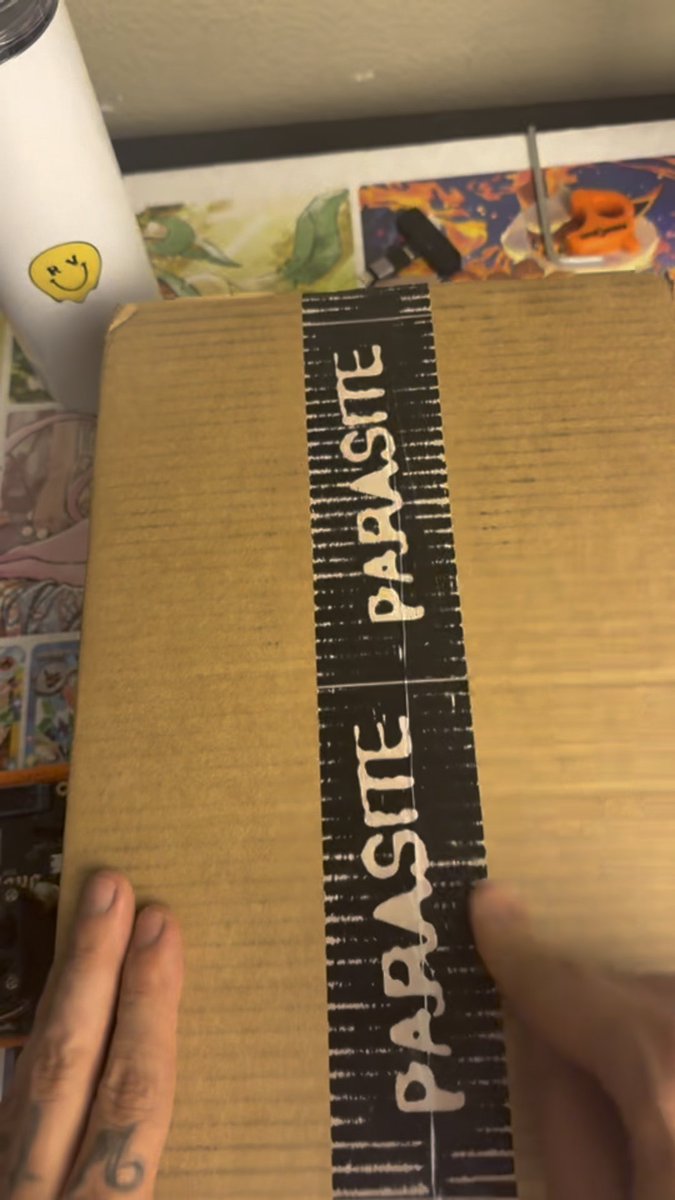 A cardboard box sealed with black "Parasite Paste" tape is being unboxed by two hands, with a white tumbler featuring a smiley face sticker visible in the background, corresponding to the unboxing of the @Parasite_wtf crypto miner mentioned in the caption.