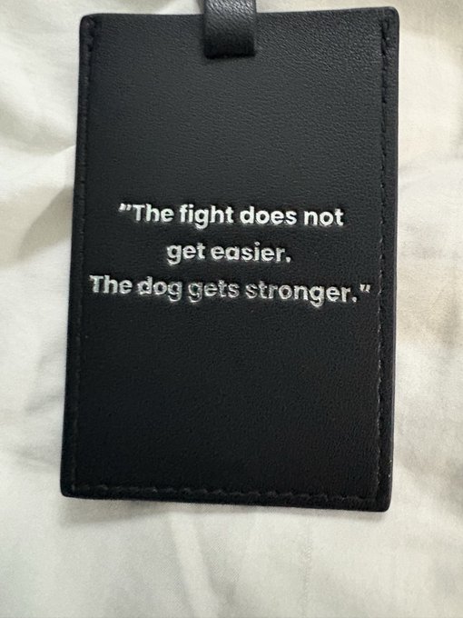 A black leather luggage tag embroidered with the quote 'The fight does not get easier. The dog gets stronger.'