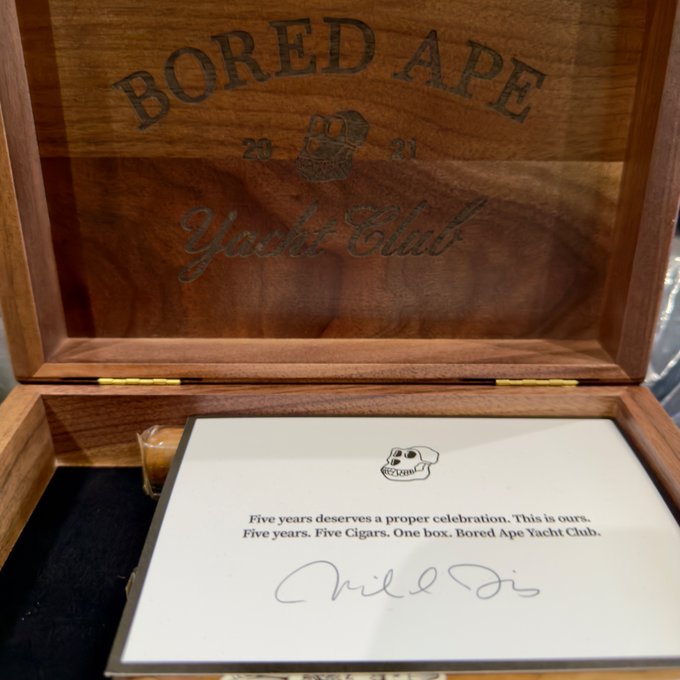 An open wooden cigar box engraved with 'BORED APE YACHT CLUB 2021' contains a card reading 'Five years. Five Cigars. One box. Bored Ape Yacht Club.' with a signature.