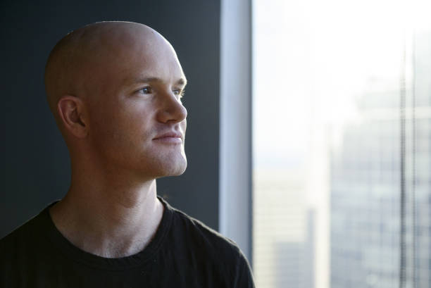 Portrait of a bald man in a black shirt looking thoughtfully out a window toward a cityscape.