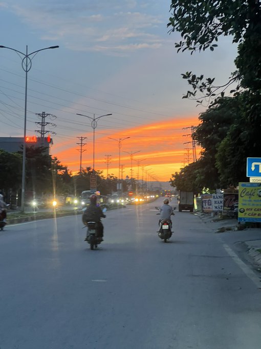 A street scene at sunset featuring motorcyclists riding down a road with an orange and pink sky, streetlights, power lines, and signage visible.