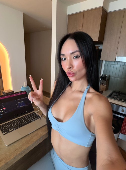A woman in a light blue matching workout set poses with a peace sign in a modern kitchen, flashing a laptop displaying trading charts. The casual "Happy Sunday" post pairs the candid selfie with a playful plea for the community to recommend high-return (100x) crypto or stock picks.