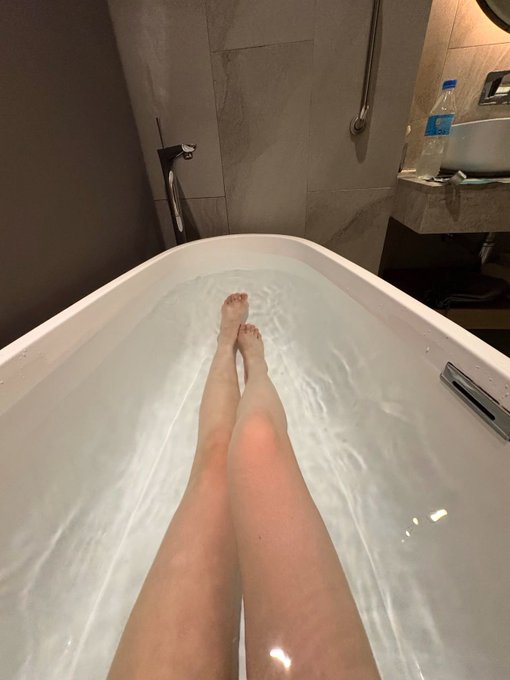 A first-person perspective photo of someone relaxing with their legs extended in a bathtub filled with water in what appears to be a hotel bathroom.