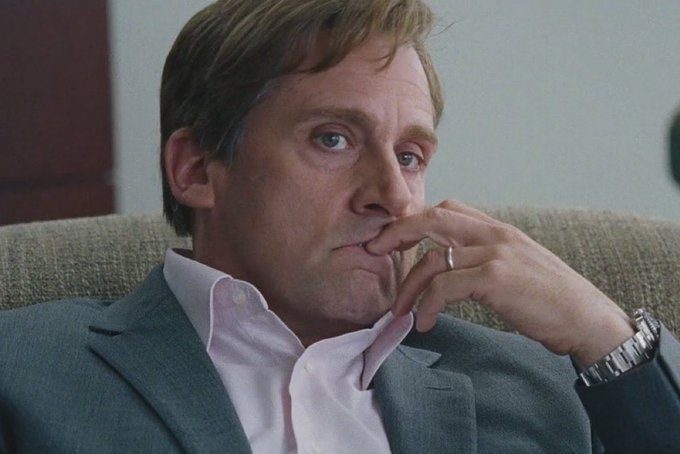 A close-up portrait of a man in a grey suit and pink shirt sitting on a couch with his hand resting on his chin, appearing deep in thought.