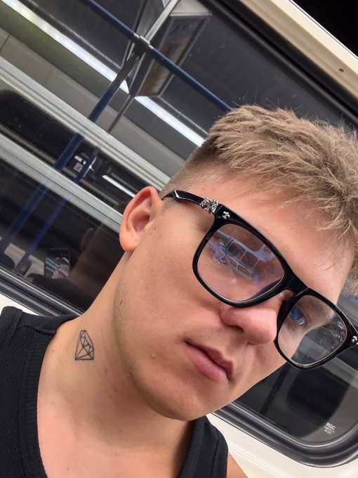 A close-up selfie of a young blonde man wearing black glasses, with a small diamond tattoo on his neck, taken inside a subway train.