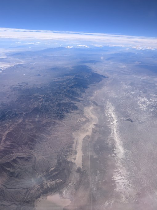 An aerial photograph captures a vast desert landscape featuring rugged mountain ranges, dry valleys, and scattered cloud cover beneath a clear blue sky.
