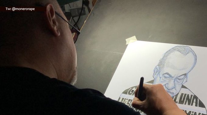 An artist with glasses is shown from behind, sketching a detailed portrait of an older man on paper taped to a board, with the partial text "LUNA" and what appears to be a football helmet visible in the drawing. The handle "@monerorapa" is displayed in the top left corner of the image.