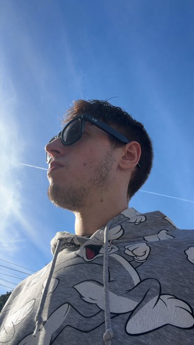 A low-angle selfie of a man with short brown hair and stubble wearing dark sunglasses and a grey Bugs Bunny hoodie, photographed against a bright blue sky with wispy clouds.