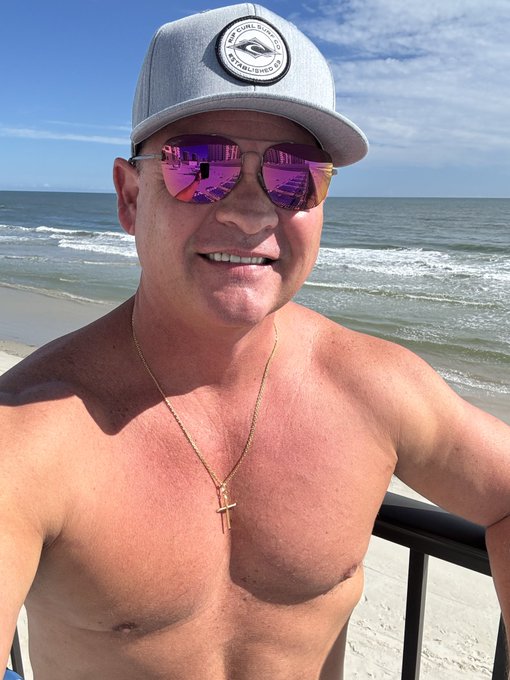 A shirtless, tanned man wearing a grey baseball cap and purple reflective sunglasses, taking a selfie on a balcony overlooking the beach.