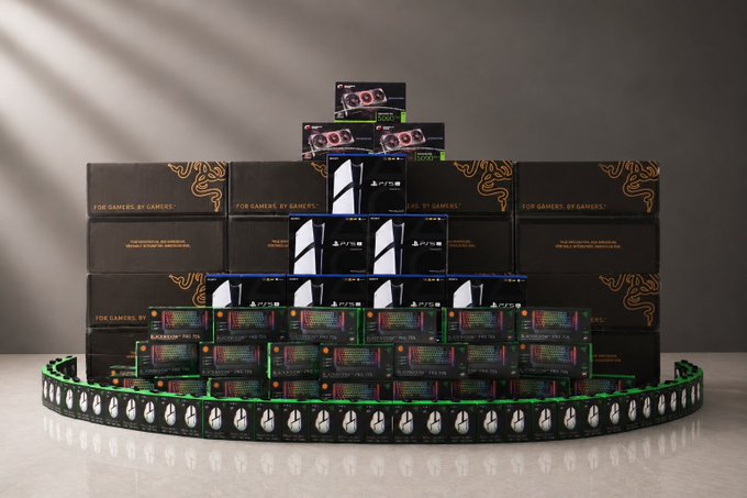 A pyramid-shaped stack of gaming hardware boxes, including PlayStation 5 and ASUS components with Razer branding, sits on a curved track illuminated with green and white lights.