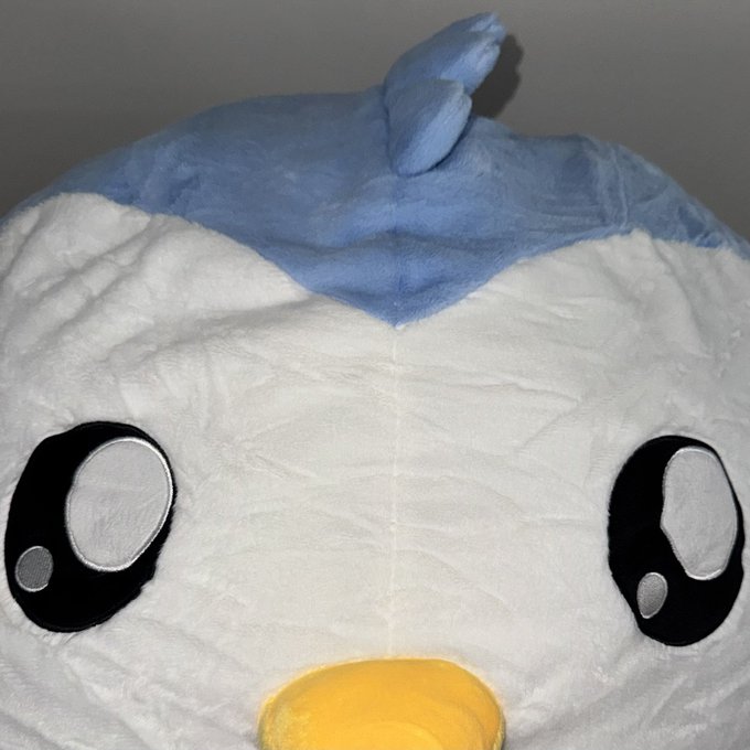 A close-up of a plush toy featuring a white face, large black eyes, a yellow beak, and a blue tuft of hair on its head.
