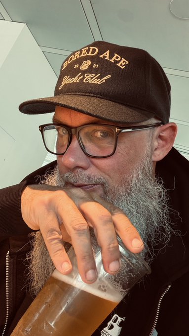 The image features a man with glasses, a beard, and a mustache wearing a black baseball cap that says "Adored Apes" on it. He has his hand resting on his face while holding a glass of beer. The background appears to be a white wall or a window, but the focus is on the man in the center of the image.

The caption provided for this image reads: "This ape is heading back to NYC and looking forward to meet my fellow New York apes and web3 guys for the next 4 weeks."