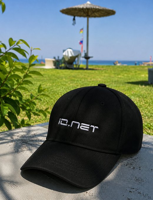 A black baseball cap with white "ID.NET" embroidery sits on a concrete surface, with a beach scene featuring a thatched umbrella, flags, and the ocean visible in the background.