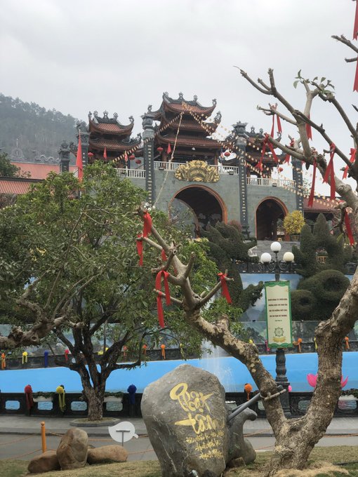 A traditional Asian temple or shrine with ornate roof details, decorative arches, and red ribbons tied to trees in the foreground, with a stone marker bearing gold lettering.
