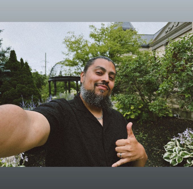 A man with a beard wearing a black shirt takes a selfie in a lush garden, giving a "shaka" hand sign while the background features greenery and a gazebo, complementing the post's confident message about his ethos_network profile.