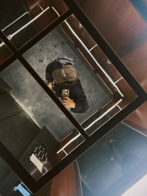 An overhead shot looking down into a mirror reflection, showing a person wearing a cap and holding a phone to take a selfie in what appears to be a stairwell or elevator.