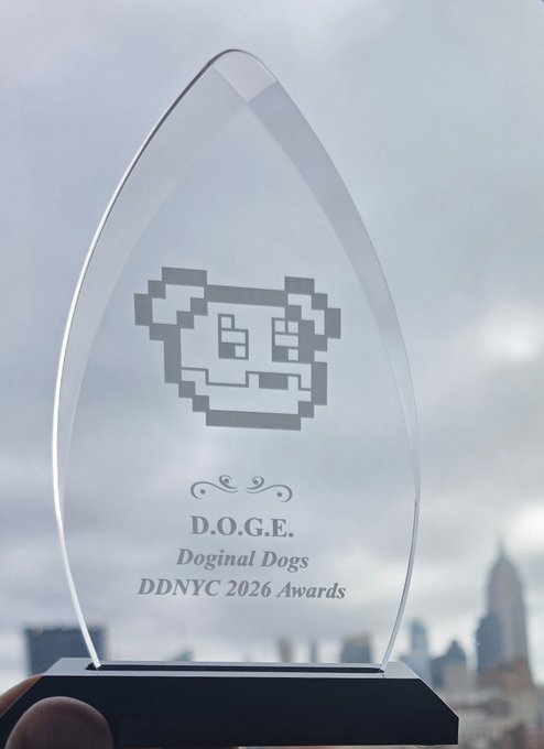 A clear glass award trophy featuring the Dogecoin pixelated dog logo engraved on it, with text reading 'D.O.G.E. Doginal Dogs DDNYC 2026 Awards' held up against a city skyline.