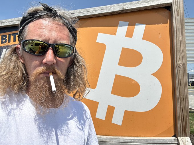 A long-haired, bearded man in sunglasses smokes a cigarette in front of an orange sign featuring the Bitcoin "₿" symbol, with partial "BITC..." text visible at the top, visually reinforcing his caption about closing down Bitcoin ATM locations after 13 years in the industry.