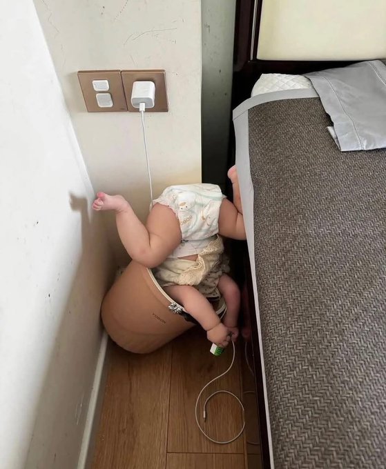 A baby in a diaper squirms in a narrow gap between a bed and wall, charging cable in hand, fittingly illustrating the chaotic, cramped effort of "doing this on Neo" — likely a meme referencing the struggle of navigating tight spaces or awkward situations.