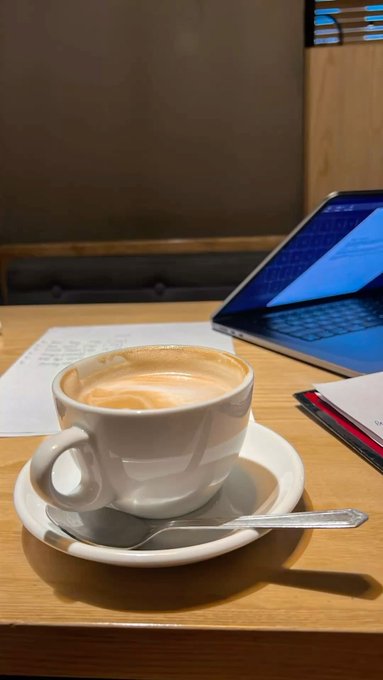 A white ceramic cup of cappuccino with latte art sits on a wooden table next to an open laptop with a blue screen, with papers visible in the background.
