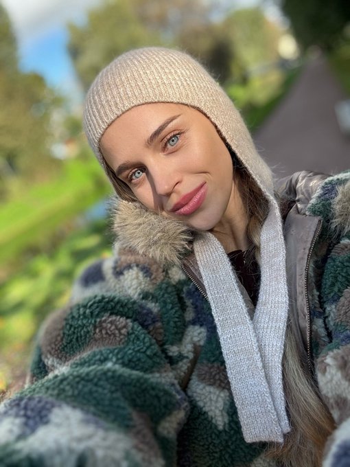 A close-up selfie of a woman with striking blue eyes, wearing a beige knit hooded scarf and a patterned fleece jacket, standing outdoors.