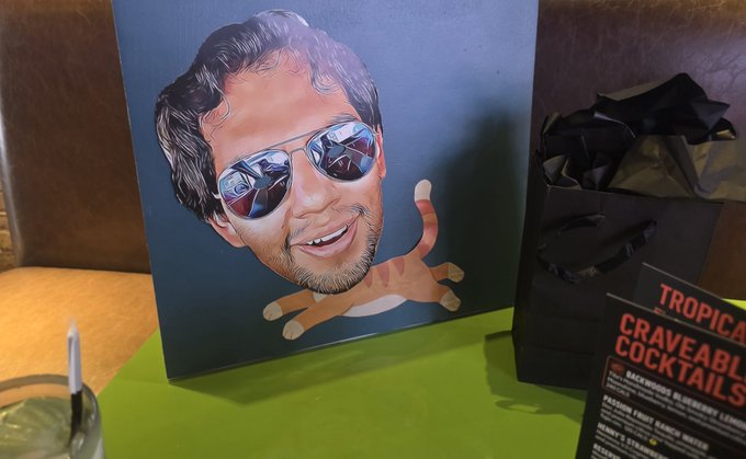 A humorous painting on a blue canvas shows a man's head with aviator sunglasses photoshopped onto the body of an orange tabby cat, displayed next to a black gift bag and a menu for "Craveable Cocktails" from "Tropical".