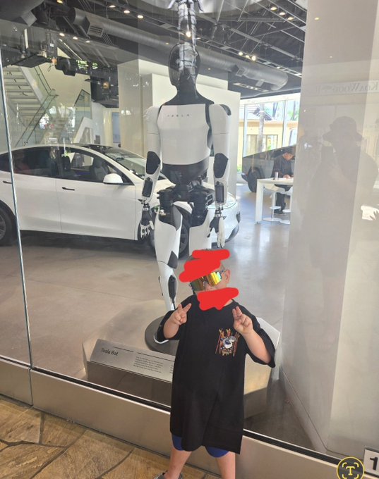 A child poses with peace signs in front of a Tesla Optimus humanoid robot and a white car at what appears to be a Tesla showroom, with a plaque reading "Tesla Bot" visible. The image aligns with the post's caption, "little bluntz wants an $optimus @Optimus_RH," showing the child's fascination with the Tesla Optimus robot and tying into the $Optimus ticker mentioned.