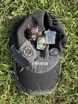 A black beanie hat with 'solana' text embroidered on it, placed on grass with small collectible figurines (including a devil) resting on top.