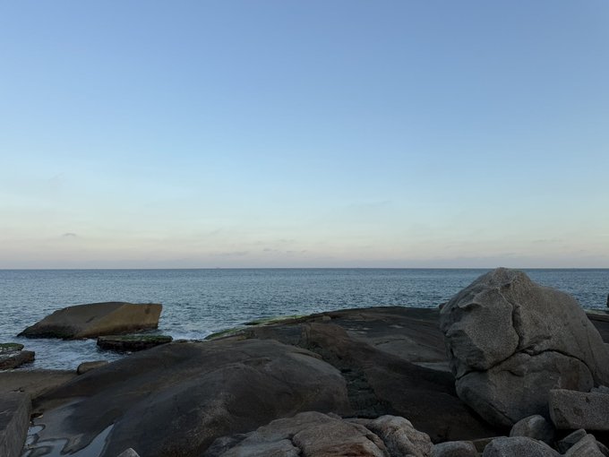 A calm seascape with rocky shoreline in the foreground under a soft blue sky at dusk or dawn. The serene coastal scene mirrors the contemplative, patient tone of the post, reflecting the waiting-and-watching sentiment about the market being "stale" with anticipation for the next big "golden dog" opportunity.