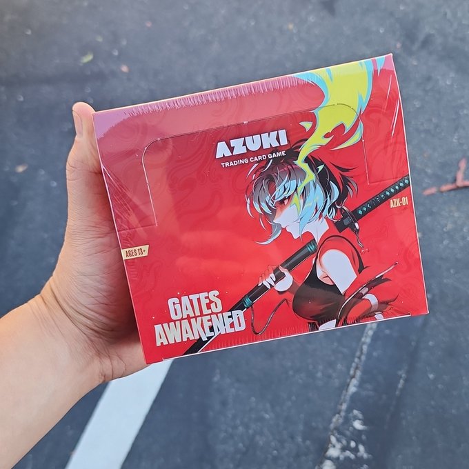 A hand holds a sealed booster box of the "AZUKI TRADING CARD GAME" titled "GATES AWAKENED" (set AZK-01, ages 13+), featuring anime-style artwork of a sword-wielding character against a vibrant red background—matching the post about purchasing the new game at a store.