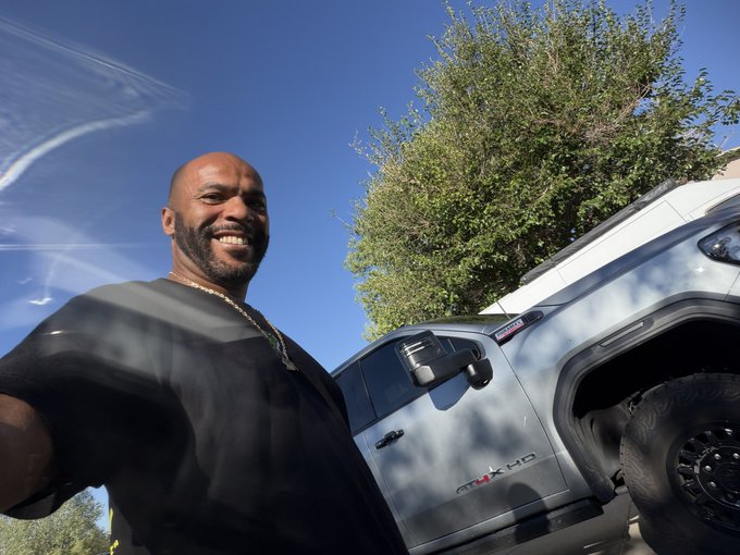 A smiling, bald, bearded man wearing a black shirt and gold chain stands in front of a white Ram TRX pickup truck outdoors under a blue sky.