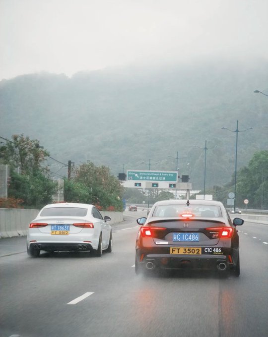 The image shows two cars driving down a road, with one car being white and the other black. The white car has its brake lights on, indicating that it might be slowing down or stopping. In the background, there are mountains visible behind the vehicles, adding to the scenic nature of the scene. The post in the image reads "粤车南下进港", which could possibly be a location name or a reference to an event related to the cars driving down the road.