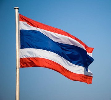 The flag of Thailand, featuring horizontal red, white, and blue stripes, waves on a flagpole against a clear blue sky.