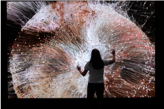A person stands silhouetted in front of a large screen displaying a colorful, abstract visualization resembling a neural network or data web.
