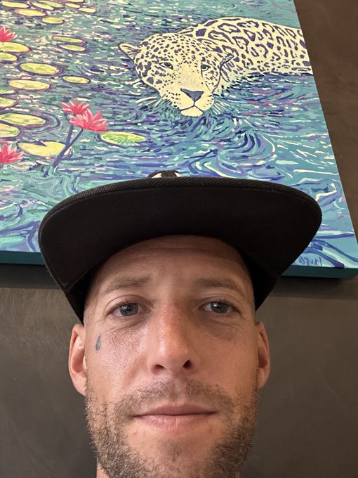 A selfie of a man wearing a black cap in front of a colorful painting featuring a leopard swimming through water with lily pads and flowers.