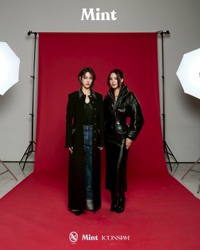 Two women pose together in a professional photo studio against a red backdrop, one in a long black coat over jeans and the other in a black leather jacket and pants, with 'Mint' at the top and 'Mint ICONSiam' at the bottom.