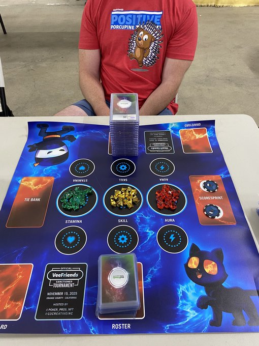 A person in a red 'POSITIVE PORCUPINE' t-shirt sits at a gaming mat labeled 'Veefriends Spirits Tournament NOVEMBER 15, 2025 ORANGE COUNTY, CALIFORNIA', with stats for Primals, Tiers, and Traits displayed.