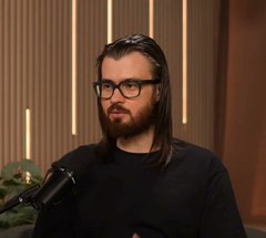 A man with long dark hair, a beard, and glasses sits in a studio setting, wearing a dark sweater near a microphone.